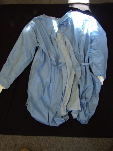 A BLUE SURGICAL GOWN FOR USE IN HOSPITALS. DESIGNED TO COVER THE BODY FROM THE SHOULDERS TO JUST BELOW THE KNEE. WITH A COMPLETE OPENING AT THE BACK WITH VELCRO FASTENING AT THE NECK AND TIE FASTENING AT THE WAIST. WITH LONG SLEEVES WITH WHITE KNITTED ELASTICATED CUFFS.  MADE FROM NON-WOVEN 55 PER CENT CELLULOSE FIBRES (PAPER) AND 45 PER CENT POLYESTER FIBRES. FLUID REPELLENT. DISPOSABLE AFTER USE.
