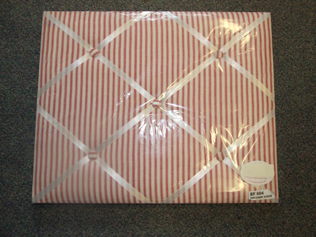 A RECTANGULAR WOODEN BOARD COVERED WITH PADDED TEXTILE. RIBBONS WHICH ARE HELD IN PLACE BY BUTTONS CRISS-CROSS OVER THE TEXTILE. NOTES AND MEMOS CAN BE SLID UNDERNEATH THE RIBBONS OR PINNED TO THE TEXTILE, SUCH THAT IT CAN BE USED AS A MESSAGE BOARD. WITH METAL FIXTURE ON THE BACK FOR HANGING ON A WALL