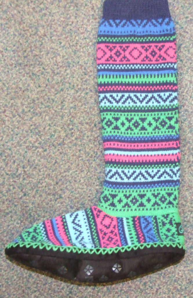 SLIPPER SOCK WITH KNITTED WOLLEN UPPER AND MICROFIBER OUTSOLE. THE SURFACE AREA IN CONTACT WITH THE GROUND IS THE SOLE.THE SOLE IS  BLACK IN COLOUR WITH A LOGO. WITH A RIBBED TOP. WITH A DESIGN WITH GREEN, BLUE, PINK COLOURS. FOR INDOOR USE. THE SOLE MEASURES 27 CM. THE PART COVERING THE LEG MEASURES 30 CM