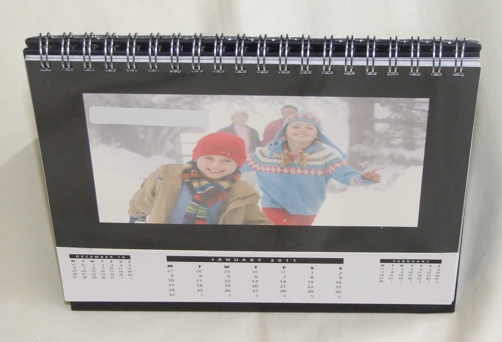 A DESKTOP CALENDAR WITH SELF LAMINATING PEAL AND SEAL WINDOWS TO SHOW PHOTOGRAPHS.  CALENDAR IS MADE OF CARDBOARD WITH PLASTIC PEAL AND SEAL FRAME AND METAL SPIRAL BINGING.  HOLDS 12 6 X 4 INCH PHOTOGRAPHS WHICH ARE PLACED UNDER A SELF ADHESIVE BLACK FRAME. THE FRONT OF EACH PAGE SHOWS A MONTH-TO-VIEW FOR THE CURRENT, PREVIOUS AND NEXT MONTH; THE BACK OF EACH PAGE SHOWS A MONTH TO VIEW WITH SPACES TO ENTER INFORMATION.  INCORPORATES A STAND OF BLACK CARDBOARD.  PACKAGED IN A HANGING PLASTIC BAG FOR RETAIL SALE.