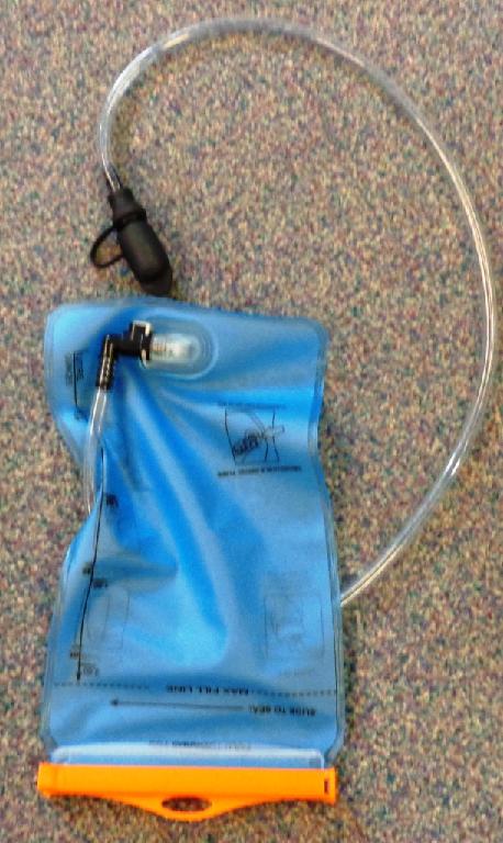 A BLUE TPU PLASTIC WATER BLADDER/WATER RESERVOIR. PRINTED WITH MEASUREMENTS ON ONE SIDE IN LITRES AND OUNCES AND INSTRUCTIONS FOR USE ON OTHER. WITH A FOLDING CLOSURE AT TOP SECURED WITH A RIGID PLASTIC BAR THAT SLIDES INTO PLACE. WITH REMOVABLE DRINKING TUBE ATTACHED BY A PUSH FITTING ON ONE END AND A STOPPER/CAP ON THE OTHER. MEASURING APPROXIMATELY 31CM X 18CM CLOSED.