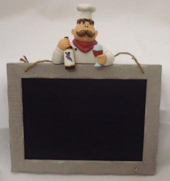 A WOODEN MENU BOARD WITH A POLYRESIN FIGURE OF A CHEF  ON TOP. THE CHEF HAS A MOUSTACHE IS WEARING A HAT AND IS HOLDING A BOTTLE OF DRINK IN ONE HAND AND A GLASS IN THE OTHER. THE FIGURE IS MADE FROM MORE THAN 50% STONE AND THE BALANCE IS PLASTIC RESIN . THE BOARD IS PAINTED MATT BLACK AND IS USED WITH CHALK.