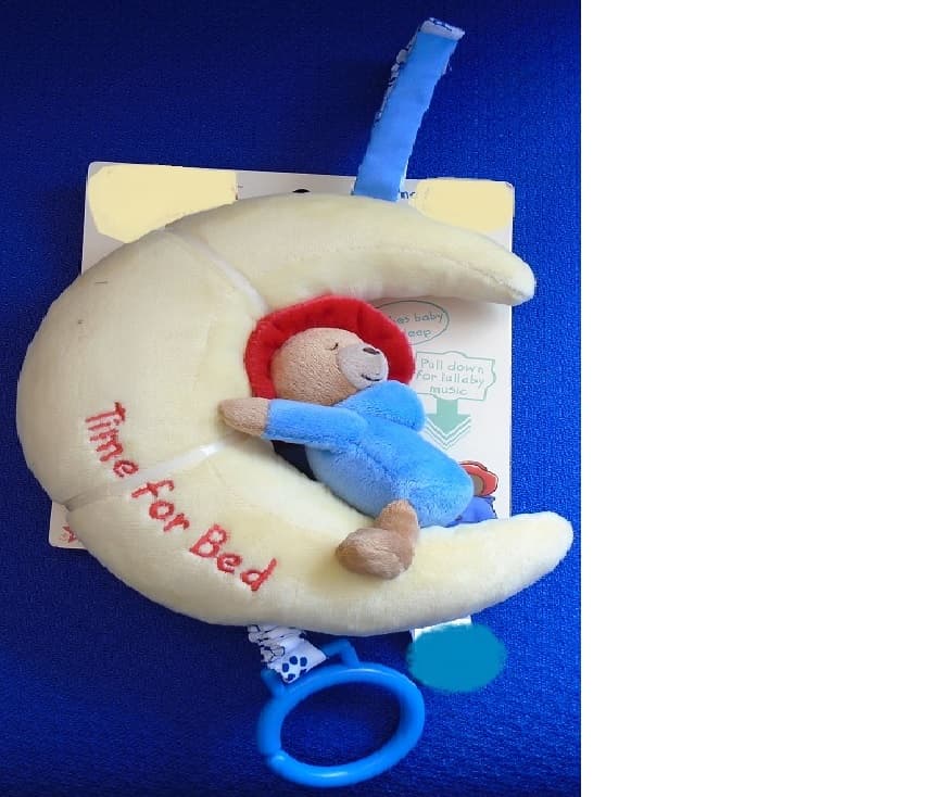 TIME FOR BED, A STUFFED MOON SHAPE WITH A WELL KNOWN CARTOON/FILM CHARACTER BEAR ATTACHED TO IT, WITH INTERGRATED MUSIC BOX. PULL THE CORD AT THE BOTTOM OF THE MOON AND IT PLAYS A LULLABY, SOOTHING THE BABY TO SLEEP. THE MOON AND BEAR ARE STUFFED IN A PLUSH MATERIAL, THE MOON IS YELLOW WITH RED EMBROIDERY SAYING TIME FOR BED. THE BEAR HAS A BLUE BODY REPRESENTING A BLUE COAT WITH A RED HAT. THERE IS A CONTRASTING BLUE HANGING STRIP AT THE TOP OF THE MOON FOR HANGING ON/OVER A COT. MEASURING APPROXIMATELY 16 ½ CM IN HEIGHT. FOR AGE FROM BIRTH. PACKAGED ON A BRIGHTLY COLOURED CARDBOARD BACKING WITH HANGING FACILITIES.