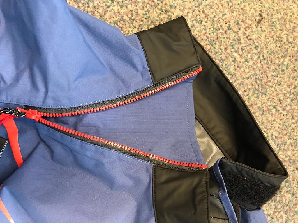 WOVEN GARMENT IN A BLUE COLOUR WITH BLACK PANELS TO THE UNDERARMS.  THERE IS A RED PRINTED STRIPE ABOVE THE BLACK PANEL.
A PARTIAL RED ZIP WHICH EXTENDS DIAGONALLY ACROSS THE CHEST TO RIGHT SHOULDER AND FINISHES AT THE ROUND NECKLINE.  NO OPENING IS CREATED WHEN UNZIPPED AS THERE IS A TEXTILE PANEL BENEATH WHICH ACTS TO INCREASE THE SIZE OF THE NECKLINE.  ATTACHED TO THE ZIPPER IS A BLACK PULL CORD WITH WHITE STRIPES AND RED TIPS.
THERE ARE ALSO VELCRO FASTENINGS TO THE NECKLINE WHICH ALLOW TIGHTENING OR LOOSENING.
THE GARMENT IS LONG SLEEVED WITH CUFFS OF PLASTIC WHICH HAVE A VELCRO AND ELASTIC FASTENING SYSTEM.
BRANDING TO THE MIDDLE CHEST AND BACK OF NECK AREA.
THERE IS VELCRO TIGHTENING TO THE BASE OF THE GARMENT.  THE BASE IS MADE FROM NEOPRENE RUBBER (INSIDE) WITH A KNITTED TEXTILE (OUTER SIDE) ON ONE SURFACE ONLY.
THE MAJORITY OF THE GARMENT IS MADE OF A WOVEN TEXTILE WITH A VISIBLE NON CELLULAR HYDROPHILIC POLYURETHANE (PU) PLASTIC ON THE INNER SURFACE.  MADE OF 100% NYLON.
THE PACKAGING IS A PLASTIC BAG WITH A STICKER TO IDENTIFY THE GARMENT ENCLOSED