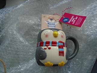 A PENGUIN SHAPED STONEWARE MUG. THE PENGUIN IS A GREY COLOUR WITH A WHITE BREAST, YELLOW BEAK AND FEET, AND A RED AND BLUE SCARF. PACKAGED WITH A SACHET OF HOT CHOCOLATE FLAVOUR DRINK POWDER IN A SEALED CARDBOARD BOX AND A BAG OF WHITE MINI MARSHMALLOWS IN A SEALED PLASTIC BAG. THE HOT CHOCOLATE AND MARSHMALLOWS ARE CONSIDERED TO BE NEGLIGIBLE ARTICLES. THE ITEMS ARE PACKAGED TOGETHER FOR RETAIL SALE WITH A PLASTIC SURROUND ENCASING THE ITEMS.