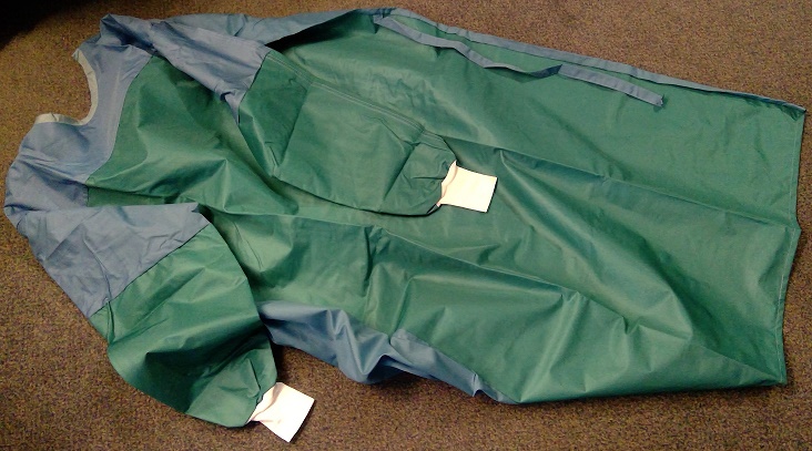 A KNITTED HIGH PROTECTION SURGICAL GOWN MADE UP OF A TRILAMINATE TEXTILE IE: THREE LAYERS, POLYESTER, POLYURETHANE AND POLYESTER. CAN BE USED FOR SURGICAL PROCEDURES WITH HIGH LEVELS OF FLUIDS. 
THE FRONT PANEL AND SLEEVES CONSISTS OF THREE LAYERS MADE UP OF TWO OUTER LAYERS BEING A KNITTED POLYESTER TEXTILE (GREEN) AND WITH A CENTRAL LAYER OF POLYURETHANE PLASTIC (WHITE) WHICH IS VISIBLE IN CROSS SECTION.
THE BACK PANEL AND TOP PART OF THE SLEEVES ARE MADE UP OF WOVEN MICROFIBRE POLYESTER FABRIC WITH A WHITE KNITTED CUFF AT THE BOTTOM OF EACH SLEEVE. 
THE MINT GREEN NECK COLLAR INDICATES IT IS A LARGE SIZE GOWN. IT HAS A COMPLETE OPENING AT THE BACK WITH  A TIE BELT FASTENING AND FOUR ADJUSTABLE PRESSTUDS AT THE BACK OF THE COLLAR.