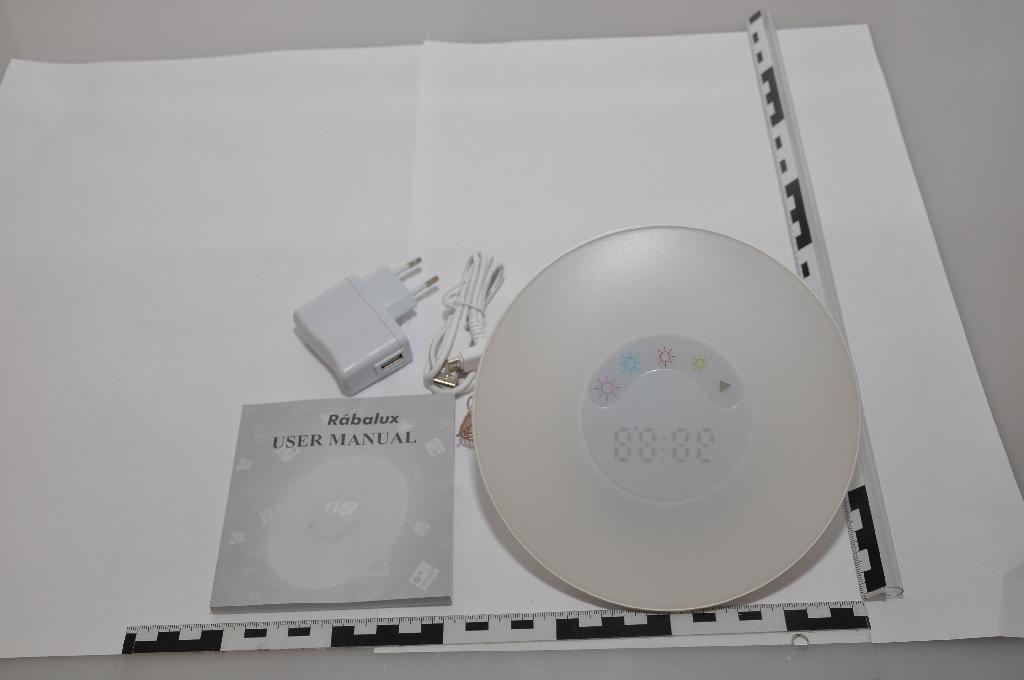 A termék lámpatest ébresztőóra és FM rádió funkciókkal, valamint hangulatfénnyel. A minta egy fehér színű műanyag asztali óra. 17 cm átmérőjű kör alakú; előlapján matt fehér áttetsző műanyag borítás látható, közepén egy kb. 7 cm átmérőjű körben a digitális kijelző, alatta 5 érintőgomb: 1 üzemmódkiválasztó és 4 fényerőszabályzó/színkiválasztó. Az óra hátlapja fényes fehér, nem áttetsző, kidomborodó; itt látható 4 nyomógomb: egy „set” feliratú, két hangerőszabályzó/állomáskereső, 1 „radio” feliratú. Továbbá a hátlapon van még sok apró, kb. 1 mm átmérőjű kör alakú lyuk a hangszórónak, egy óratámaszték, egy vékony antennakábel, egy fedéllel eltakart elemdoboz 2 db AAA elemnek, és egy 5V-os mini USB bemeneti csatlakozóaljzat. A dobozban található még egy fehér színű hálózati kábel, egyik végén 2.0-s USB-A, másik végén 2.0-s mikro USB-B csatlakozóval, valamint a hozzá tartozó USB-A portos hálózati adapter.
A használati útmutató alapján a termék „rádiós ébresztőóra hangulatvilágítással”, „nem világításcélú lámpatest”.
Az ügyfél nyilatkozata szerint a termék anyaga műanyag, mely egyezik a vizsgálati eredményekkel.
Egyéb információk: A termék (1,8 W) és RGB (2,7 W) LED izzókat tartalmaz, elemről és külső áramforrásról is működtethető. A termék kiskereskedelmi forgalom számára kiszerelt.