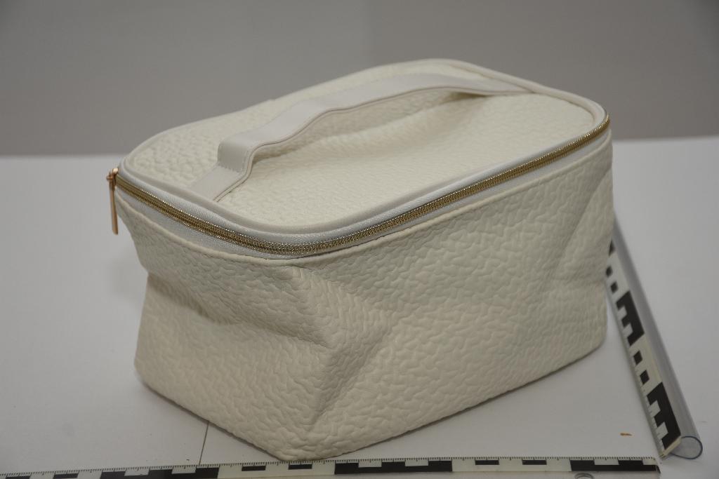 A minta tört fehér színű, nyomott mintával bordázott, sarkain lekerekített, téglalap alapú, 21,5 cm hosszú, 15,5 cm széles, 13,5 cm magas piperetáska szintetikus külső felülettel.  A táska teteje körben húzózárral nyitható. A táska tetején fehér színű, hosszában mindkét oldalon rávarrt 2,2 cm széles pántból készült fül található. A minta alja és a hátuljának közepén 13,2 cm széles sávban tört fehér színű, sima felületű anyagból készült. A táska külső felülete bordás tapintású műanyag. A minta fehér színű, szőtt béléssel ellátott. A minta külső felületének belső oldala (hordozó) kötött/hurkolt kelme. A minta bélése és a külső felület között, fehér színű nemszőtt textília réteg található.
A minta külső felülete főként poli(vinil-klorid) és poliészter műanyag.