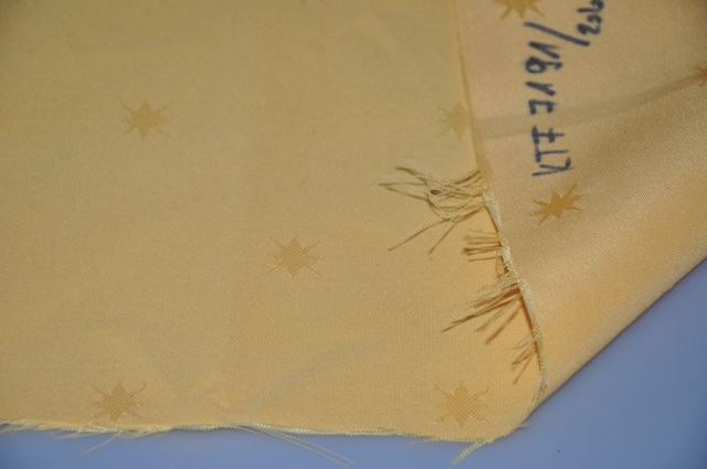 A termék jacquard szövésű textil. A minta csomagolás nélkül érkezett a laboratóriumba. Analitikai vizsgálatok: Érzékszervi vizsgálat: 
A minta sárga színű, téglalap alakú, önmagában csillagokkal díszített szövet, mind a négy oldalán vágott széllel. A mintán a „Republika Slovenska, Ljubljana” a bélyegző lenyomata található. Sem a színoldalán, sem a hátoldalán szabad szemmel bevonat, impregnálás nem látható. Beküldött minta  mérete: Szélesség: 41 cm; Hosszúság: 42,5 cm. Kelmeképzés: szőtt, jacquard szövésű; láncfonal: végtelen, terjedelmesített, sodrott, egyágú; vetülékfonal: végtelen, nem terjedelmesített, nem sodrott, egyágú. Mikroszkópi vizsgálat (8x nagyítás): a felületen impregnáló szer látható. Kölcsönhatás vízzel: vízlepergető tulajdonságú; vízben áztatva mindkét oldala nedvesedik, nem vízhatlan. Nyersanyag összetétel (MSZ 13560-2:1989): poliészter.
Szakítóvizsgálat (MSZ EN ISO 2062:1999): Láncfonal szakító-szilárdsága: 29 cN/tex, Vetülékfonal szakítószilárdsága: 31 cN/tex.
A XI. Áruosztály Megjegyzések  (6) pontja alapján a poliészter szálakból készült lánc és vetülékfonal nem nagy szakítószilárdságú.
Terjedelmesített fonalarány meghatározása (25/2005. (IV. 29.) GKM rendelet, kézi szétválasztás): Terjedelmesített fonalak aránya: 59,7 %
Az elvégzett vizsgálatok alapján a minta végtelen poliészter szálakból jacquard szövéssel készült. A terjedelmesített fonalak aránya: 59,7 %. Sem a lánc, sem a vetülékfonal nem nagy szakítószilárdságú.