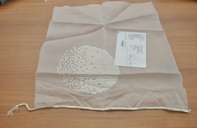 A jogosult nyilatkozata alapján a termék 100 % poliészterből készült, gépi hímzésű méteráru, ólomzsinórral rendelkezik; a termék minden tekintetben megegyezik a VPVI 6698-1/2010. számú Vizsgálati jegyzőkönyvben szereplő termékkel, ami alapján az alábbi jellemzőkkel rendelkezik:
A termék vajszínű, átlátszó, ráeső fényben csillogó függönydarab, melynek 3 oldala vágott, egyik oldalán interlock öltéssel szegett és az öltésházba fűzött ólomdarabokkal ellátott fonatolt textilanyaggal készült. A mintadarabon egy 19,5 cm átmérőjű kört alkotó, nem egyenletesen elhelyezkedő négyzeteket formázó vajszínű fonallal hímzett minta található.
Egyik oldalán interlock öltéssel szegett és az öltésházba fűzött ólomdarabokkal ellátott fonatolt textilanyaggal készült. Feltehetően nem kézi készítésű.
Kelmeképzés: szőtt
Alapanyag (MSZ 13560-2:1989): poliészter (szintetikus)