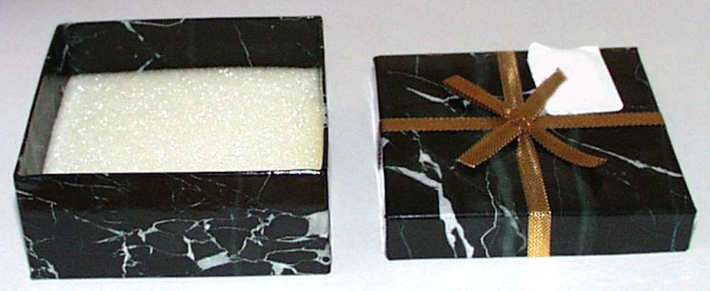 A BOX WITH LID WHICH IS SQUARED-SHAPED. IT IS MADE OF CARDBOARD AND COVERED WITH A PLASTIC, COATED, PRINTED PAPER. IT CONTAINS ONE SMALL SQUARE PIECE OF FOAM MADE OF CELLULAR PLASTIC. THE LID HAS A GOLD RIBBON TIED ACROSS IT. THE BOX IS SUITABLE FOR USE TO CONTAIN A GIFT. THE WEIGHT OF THE CARDBOARD IS APPROXIMATELY 700 G/M.SQ.