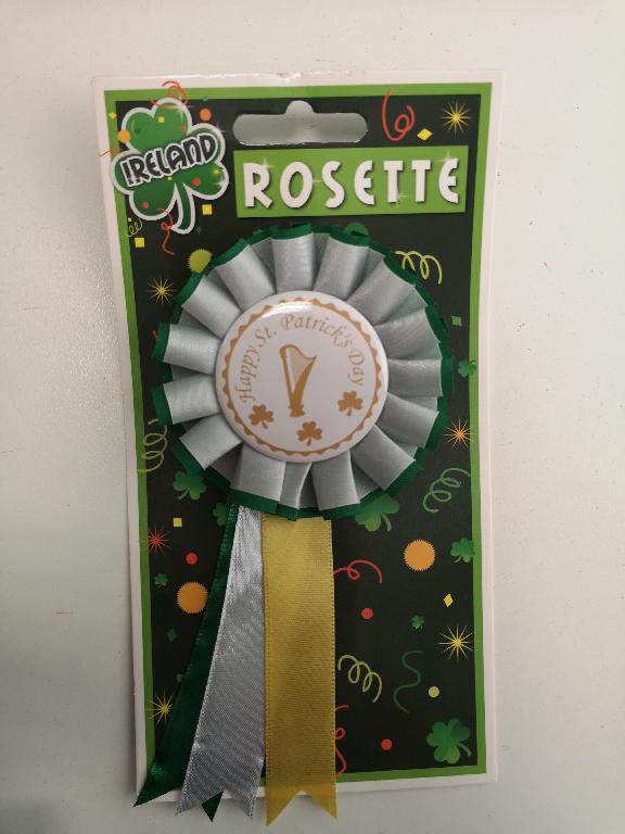 A novelty badge, circular in shape with a frill around the edge and 3 pieces of ribbon hanging down, the centre piece is edged with a gold trim and written in gold at the centre is Happy St. Patrick's Day with an image of a harp and 3 shamrocks underneath the text. There is a pin at the back with which to affix the badge to clothing.