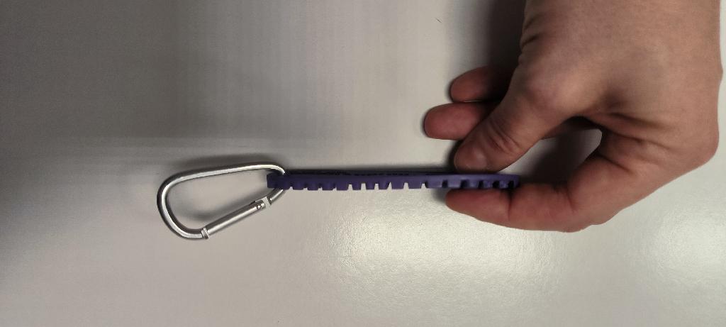 Pendaglio pubblicitario composto da un gancio in alluminio con chiusura a molla e da una parte principale in gomma vulcanizzata di colore viola, sagomata a forma di suola di calzatura. Su questa suola è presente un logo ottagonale giallo, realizzato in gomma vulcanizzata e integrato direttamente nella suola tramite pressatura e co-stampaggio, risultando parte integrante del manufatto. Secondo le informazioni fornite, le gomme per la suola e il logo vengono ottenute mescolando le materie prime, quindi ritagliate e sagomate con il peso corretto per la pressatura. I due pezzi di gomma cruda vengono inseriti da un operatore in una pressa a caldo, che unisce le due gomme. Dopo il raffreddamento, l’eccesso di materiale viene rimosso tramite una macchina rifilatrice. Il gancio in alluminio, fornito insieme al pendaglio, ha la funzione di consentire il fissaggio a borse, zaini e altri oggetti per promuovere il marchio aziendale. La gomma vulcanizzata, per dimensioni e funzione, rappresenta l’elemento principale del prodotto, mentre il gancio ha esclusivamente una funzione accessoria di fissaggio.