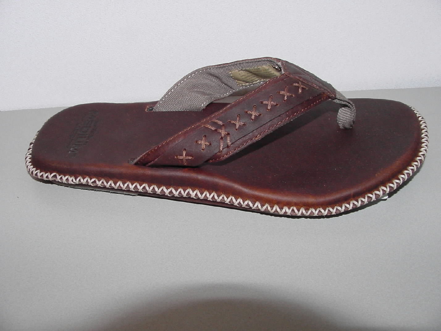 Een slipper met een bovendeel van leder en een buitenzool van rubber, niet als dames- of herenschoen te onderkennen. De slipper bestaat uit twee delen die aan de buitenzijde door een sierstiksel aanéén zijn gezet. Het bovendeel bestaat uit een V-vormige band die naast de voet in de zool is bevestigd. Op de voorzijde van de V-vormige band is een lint van textiel aangebracht die tussen de tenen in de zool is bevestigd. Op de lederen bovenband is een smallere lederen band geplaatst die met sierstiksels op de onderband is vastgezet. De buitenzool bestaat uit twee delen rubber die onder de voorvoet en hak zijn aangebracht.