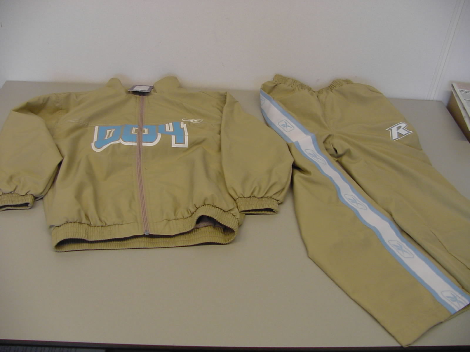 Een trainingspak vervaardigd van 100% polyester weefsel, waarvan de buitenzijde is vervaardigd van één en dezelfde stof. Het trainingsjack heeft de volgende kenmerken:
- een opstaande kraag;
- een gehele opening aan de voorzijde, voorzien van een ritssluiting;
- de uiteinden van de mouwen en de onderzijde van het jack zijn nauwsluitend door een elastische band;
- aan de voorzijde twee steekzakken;
- in de mouwen een voering van een weefsel van 100% polyester;
- in het voor- en achterpand een voering van opengewerkt breiwerk van 100% polyester;
- op het voorpand staat het logo en een decoratieve print afgebeeld.
De broek heeft de volgende kenmerken;
- nauwsluitend in de taille door middel van een elastische band;
- lange broekspijpen die aan de uiteinden zijn omgezoomd;
- op de heupen voorzien van een steekzakken;
- een decoratieve print op het linker bovenbeen;
- op de zijkant van de broekspijpen is een decoratieve band opgenaaid;
- de broekspijp is gevoerd met een opengewerkt breiwerk tot onder de knie en een dicht weefsel tot aan het uiteinde van de broekspijp.