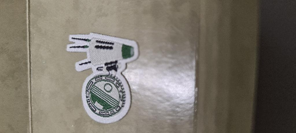 Een zogenoemde her-plakbare sticker met -volgens opgave- onder andere de volgende kenmerken:
- een embleem (badge) bestemd om op verschillende artikelen te plakken, zoals kleding, telefoon en laptop, voor het opleuken en het persoonlijker maken ervan;
- vervaardigd van geweven textiel (rPET-draad / gerecycled polyester) en een kleeflaagje;
- met ingeweven inscripties en motieven (sciencefiction);
- uit één stuk gesneden;
- niet omzoomd;
- zelfklevend;
- in verschillende vormen en afmetingen.