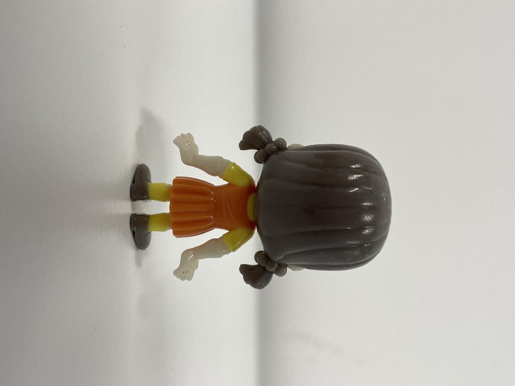 Een speelgoedfiguur met -volgens opgave- onder andere de volgende kenmerken:
- voor het vermaak van kinderen;
- een rechtop staand, niet buigzaam, meisje met 2 staartjes en een overgooier;
- vervaardigd van gerecycled kunststof;
- een hoogte van ongeveer 4 cm.

Elk figuurtje wordt blind verpakt in een bedrukte, verzegelde en ondoorzichtige papieren zak.