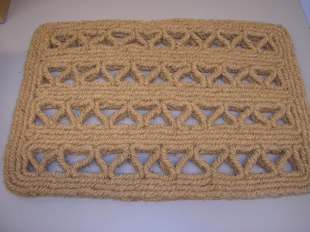 Kokosmat met een afmeting van ongeveer 41 x 58 cm. Deze mat is vervaardigd van een getwijnde kokosdraad die tot een plat koord is gevlochten. Dit koord is onderling aan elkaar verbonden tot een rechthoekige mat met over de mat verdeeld vier sier V-stroken van hetzelfde koord.