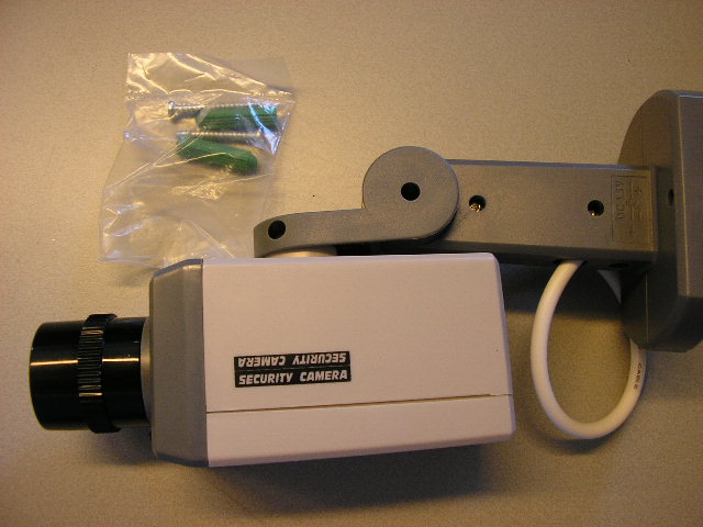 Een bewegingsmelder in de vorm een "dummy" bewakingscamera. De camera, heeft geen opnamemogelijkheid en is bedoeld als preventiemiddel, waardoor het potentiële inbrekers afschrikt. Het toestel is voorzien van de volgende kenmerken:
- een behuizing van kunststof met kabel;
- een nep lens;
- een detector of bewegingssensor, die wanneer verstoringen in haar detectiegebied worden waargenomen, de camera naar links en rechts laat bewegen; 
- een knipperende rode LED, werkend bij bovengenoemde detectie;
- een ingebouwde elektromotor, werkende op drie AAA batterijen.
De bewegingsmelder, opgemaakt voor de verkoop in het klein, verpakt in een kartonnen doos wordt tezamen geleverd met een bijbehorende muursteun en bevestigingsmateriaal.
Het apparaat is niet te onderkennen als een alarmtoestel of fopartikel.