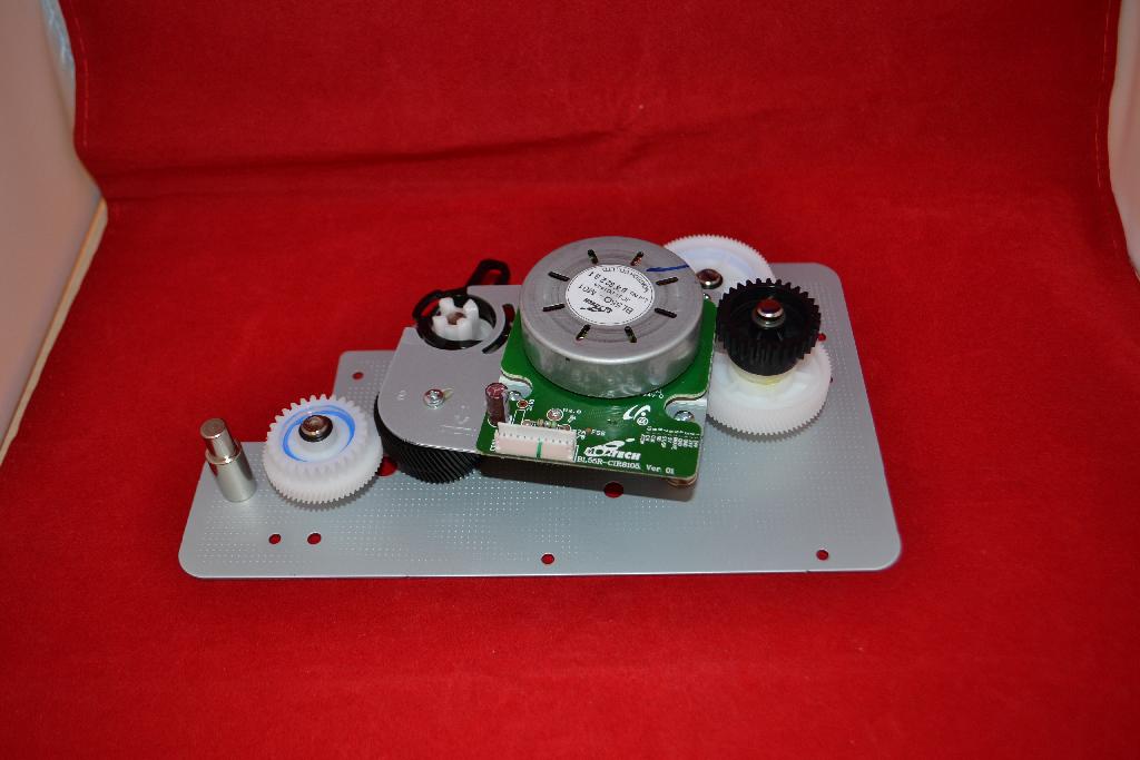 Een zogenoemde main drive, zijnde een tandwielkast, met onder meer de volgende uiterlijke en technische kenmerken:
- een metalen grondplaat, speciaal gevormd en voorzien van bevestigingsgaten;
- diverse tandwielen, vervaardigd van kunststof, in verschillende maten en met verschillende inkepingen;
- een printplaat voorzien van een aansluitpunt voor een kabel;
- een spoel;
- bestemd voor inbouw in een printer.
Het artikel heeft de functie het papier door te voeren in samenwerking met –volgens opgave- een controller.