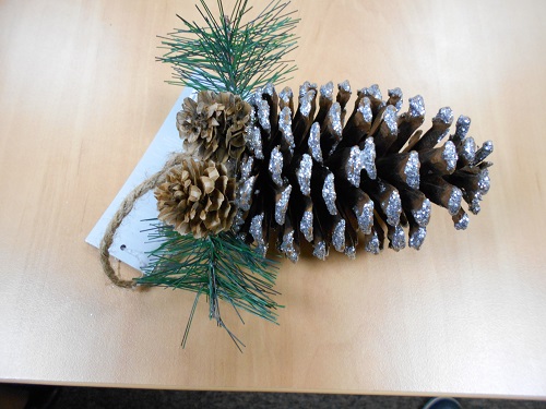 Artykuł dekoracyjny wykonany z naturalnej szyszki wielkości ok. 11 x 7 cm, wysuszonej i pokrytej częściowo farbą brokatową w kolorze srebrnym połączonej z imitacją gałązki drzewa iglastego wykonaną z drutu i z tworzywa sztucznego o długości ok. 13 cm oraz przymocowanymi do niej dwiema mniejszymi naturalnymi szyszkami wielkości ok. 3  x 3 cm z jednej strony oraz dwoma kulistymi elementami w kolorze czerwonym z tworzywa sztucznego imitującymi owoce o średnicy ok. 1,2 cm z drugiej strony. Dodatkowo  w centralnej części ozdoby u nasady szyszki umocowano pętlę ze sznurka umożliwiającą zawieszenie produktu.