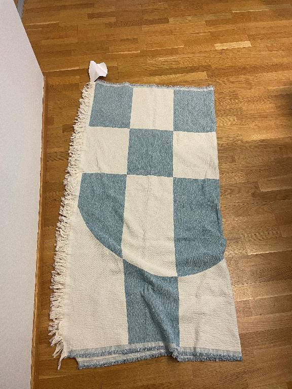 Konfektionerad artikel i form av en vävd (ej handgjord) rektangulär filt med måtten 140 x 152 cm. Filten är enligt uppgift huvudsakligen tillverkad av syntetmaterial bestående av 56 % polyester och 44 % bomull. Filten är mönstrad och avslutas med vävd kant med frans.
Filten är avsedd att svepas runt eller täcka en persons kropp för att skydda mot kyla.